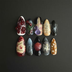 May include: A set of ten artificial nails with various designs. The designs include floral patterns, text, and a miniature portrait. Colors include red, blue, black, white, and orange. The nails are arranged on a black surface.