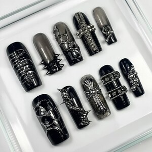 May include: A collection of ten long, coffin-shaped press-on nails. The nails are black and gray, with various metallic embellishments, including spikes, chains, and crosses. The nails are arranged on a white surface.