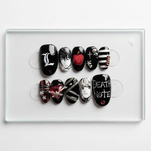 May include: A set of black and white press-on nails with anime-inspired designs. The designs include a red apple, character portraits, and the text "Death Note". Some nails feature silver chain and gem accents.