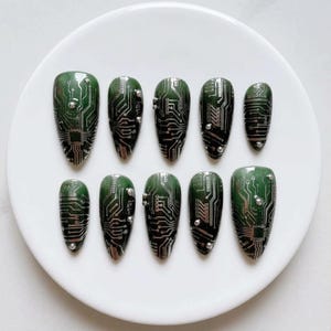 May include: A set of ten almond-shaped press-on nails featuring a dark green base with a silver circuit board pattern. Each nail is decorated with small, metallic silver studs placed along the edges and surface of the printed electronic design.