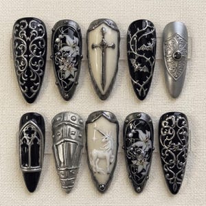 Può includere: Un set di dieci unghie finte a mandorla con intricati disegni 3D in nero, bianco e argento metallizzato. La collezione include motivi come filigrane ornate, gigli bianchi, una spada centrale, pipistrelli, un unicorno e archi gotici.