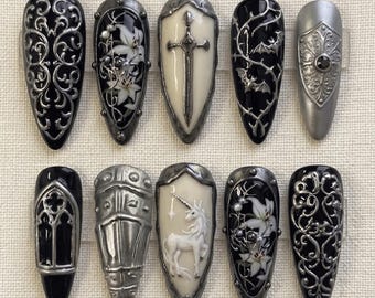 Puede incluir: Un juego de diez uñas postizas en forma de almendra con intrincados diseños en 3D en negro, blanco y plata metálica. La colección incluye motivos como filigranas ornamentadas, lirios blancos, una espada central, murciélagos y un unicornio.