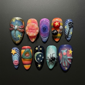 May include: A collection of ten artificial nails, each with a unique design. The nails showcase a dragonfly, the text "Welcome home!", swirling patterns, stars, a key, a spider, and a cartoon character. The colour palette includes blue, orange, purple, and black.