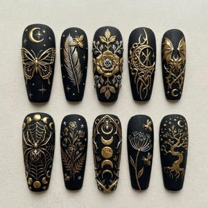 May include: A set of ten matte black press-on nails featuring raised gold and silver metallic relief designs. The motifs include a butterfly, a feather with a bee, a rose, a crescent moon, a bat, a spider web, flowers, moon phases, and a tree.