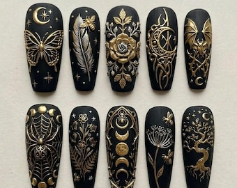 Op de afbeelding: Een set van tien matzwarte press-on nagels met verhoogde gouden en zilveren reliëfdesigns. De motieven omvatten een vlinder, een veer met een bij, een roos, een maansikkel, een vleermuis, een spinnenweb, bloemen, maanfasen en een boom.