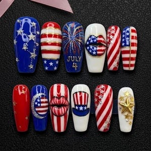 May include: A set of ten press-on nails with a patriotic theme. Designs include stars, stripes, fireworks, and the text "July 4". Colors are red, white, and blue, with gold and silver accents. The nails are arranged on a black surface.