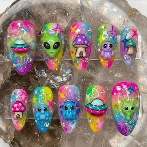 May include: A set of ten press-on nails featuring a vibrant, multi-colored cosmic background with glitter and star accents. The nails are decorated with three-dimensional charms including green alien heads, colorful mushrooms, flying saucers, and small figures.