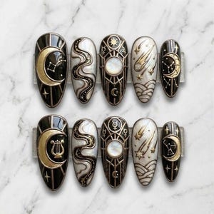 May include: A set of ten press-on nails with celestial designs. Each nail features a black base with gold and white accents, including moons, stars, and geometric patterns. The nails are oval-shaped and arranged on a clear display.