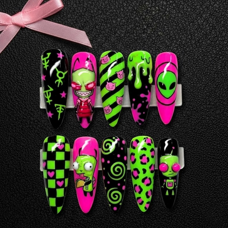 May include: A set of ten long, almond-shaped press-on nails with cartoon character designs. The nails feature black, neon green, and hot pink colors with various patterns, including aliens, pigs, and checkerboard designs. A pink ribbon is in the background.