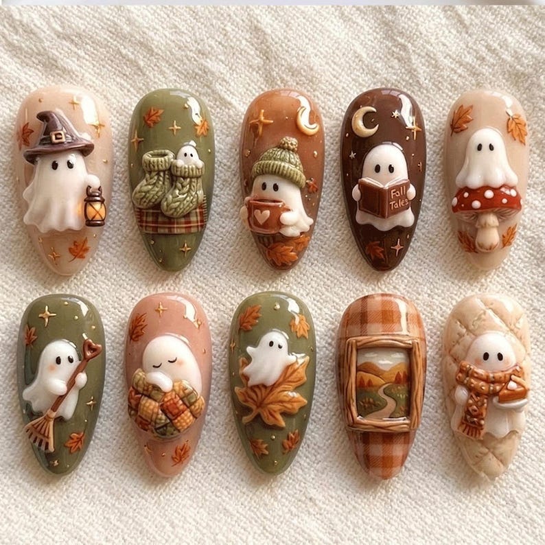 May include: A set of ten almond-shaped press-on nails featuring 3D autumn-themed designs. Each nail displays a white ghost character interacting with seasonal elements like a witch hat, lantern, knit socks, mug, book titled Fall Tales, mushroom, broom, quilt, leaves, landscape painting, and scarf.