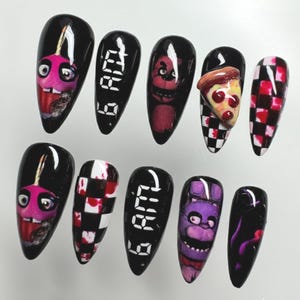 May include: A set of ten black, almond-shaped artificial nails. Each nail features a different design, including cartoon characters, checkered patterns, and the text "6 AM" and "2:48 AM". The nails are painted with a glossy finish.