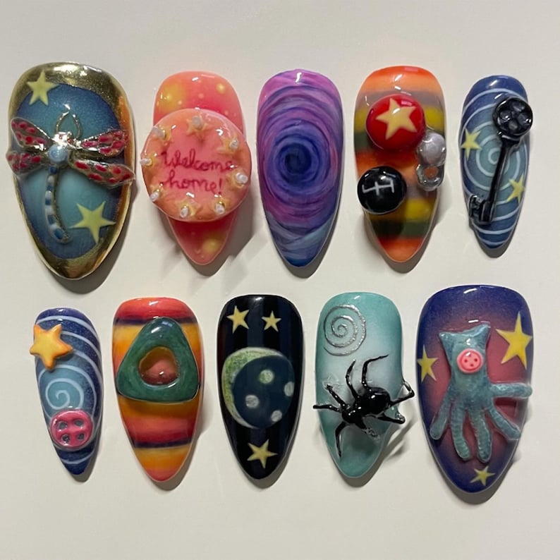 May include: A set of ten acrylic nails, each with a unique design. The nail art includes a dragonfly, a cake with the text "Welcome home!", swirling patterns, stars, a key, and a spider. The nails are painted in various colours.