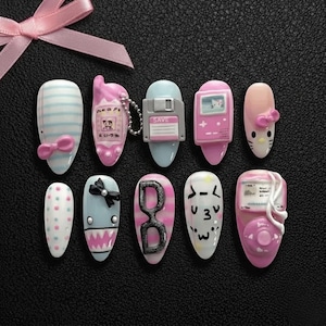 May include: A collection of ten artificial nails with various designs. The nails feature pastel colors, bows, stripes, and cartoon-like illustrations, including a floppy disk, a phone, and a video game console. A pink ribbon is in the upper left corner.