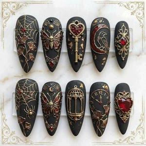 May include: A set of ten black, almond-shaped press-on nails with intricate gothic designs. Each nail features gold and red accents, including spiderwebs, butterflies, keys, and heart-shaped gems. The nails are arranged on a clear display.