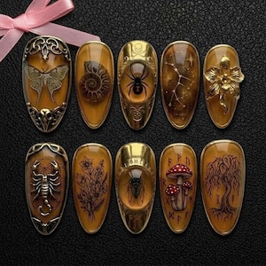 Op de afbeelding: Een set van tien sierlijke, goudkleurige nagelontwerpen. Elke nagel heeft een uniek ontwerp, waaronder een vlinder, spin, sterrenbeeld en bloemmotieven. De nagels zijn op een donkere ondergrond geplaatst.