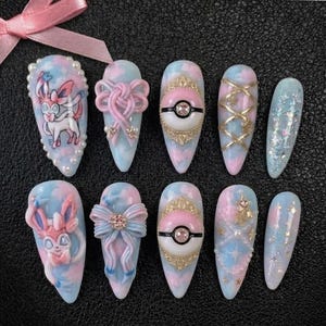 May include: A set of ten artificial nails in various designs. The nails feature a pastel blue and pink color scheme with embellishments like bows, cartoon characters, and glitter. Some nails have a Pokemon theme.