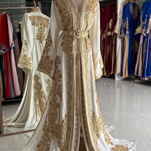Op de afbeelding: Een witte, vloerlange jurk met ingewikkelde gouden borduursels. De jurk heeft lange mouwen, een V-hals en een riem met juwelen. De jurk wordt op een mannequin gepresenteerd, waarbij het elegante ontwerp en de details worden benadrukt. Andere jurken zijn zichtbaar op de achtergrond.