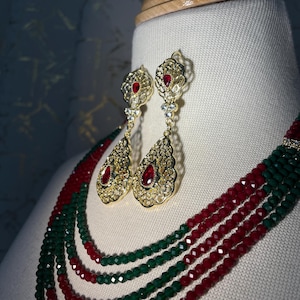May include: Gold-toned filigree earrings with teardrop-shaped pendants, each featuring a large, oval, red gemstone. The earrings are displayed with a matching necklace of red and green beads, creating a luxurious jewelry set.