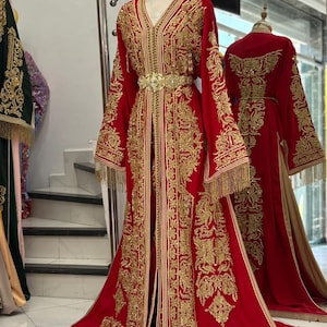 May include: A red, floor-length caftan with intricate gold embroidery and a matching gold belt. The caftan features long sleeves with gold fringe and a V-neckline. Another caftan is visible in the background.
