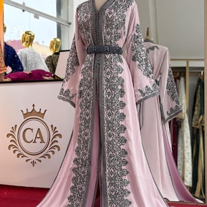May include: A light pink, floor-length gown with intricate gray embroidery and a matching belt. The gown is displayed on a mannequin, showcasing its flowing design and detailed embellishments. The gown has long sleeves and a V-neckline.