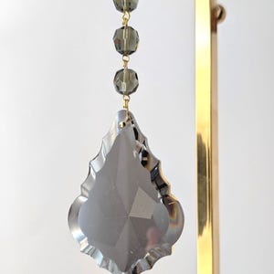 May include: A decorative chandelier crystal with a faceted, gray, teardrop-shaped pendant. Three gray, round beads are suspended above the pendant by gold-colored wire. The crystal hangs from a gold-colored metal stand against a white background.