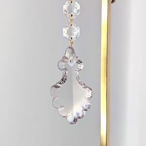 May include: Clear crystal chandelier prism with a decorative shape, suspended by gold-colored chains and a gold-colored metal frame. The prism has a scalloped edge and is designed to reflect light. The background is a neutral white.