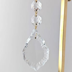 May include: Clear crystal chandelier prism with two octagonal crystals above and a large teardrop-shaped prism below. The prisms are connected with gold-colored metal links and a cream-colored ribbon. The background is a light neutral color.
