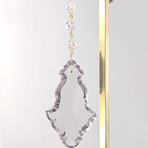 May include: A clear, decorative crystal suncatcher with a unique, ornate shape. The suncatcher is suspended by a gold chain and features three iridescent crystal beads. The top is attached to a white ribbon.