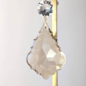 May include: A clear crystal chandelier pendant with a faceted, teardrop shape and a smaller, hexagonal crystal above it. The crystals hang from a gold-colored metal fixture.