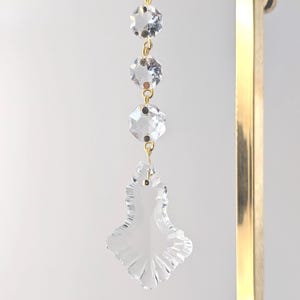 May include: A clear crystal chandelier prism with three faceted crystal beads suspended by gold-colored chains and a white ribbon. The bottom prism is leaf-shaped. A gold-colored metal support is on the right side.