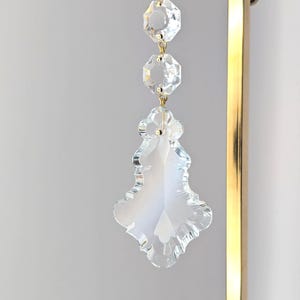 May include: A clear crystal chandelier prism with three octagonal beads and a decorative bottom piece. The prism is suspended by a white ribbon and gold-colored metal accents. The background is a light gray.