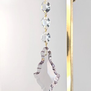 May include: A clear crystal chandelier prism with three octagonal beads and a large, decorative, teardrop-shaped pendant. The crystals are connected with gold-colored metal. The pendant has a scalloped edge and is suspended from a gold-colored metal stand.
