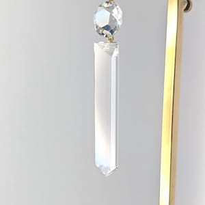May include: A clear crystal prism with a faceted, round crystal above it, suspended by a white ribbon. The prism hangs in front of a gold-colored metal stand. The crystals reflect light.