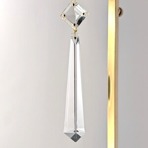 May include: A clear crystal pendant ornament featuring a diamond-shaped top piece connected to a long, tapered, multi-faceted drop. The crystal is suspended by a thin white ribbon and hangs from a vertical gold-toned metal display stand.