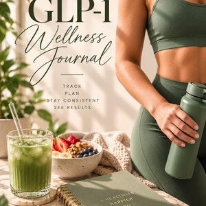 Op de afbeelding: Een saliegroen spiraalgebonden wellness-dagboek met de tekst A HEALTHIER HAPPIER YOU en een hartpictogram op de kaft. Het dagboek wordt vergezeld door een gouden pen, een glas met een groene drank en ijs, en een schaaltje met fruit.