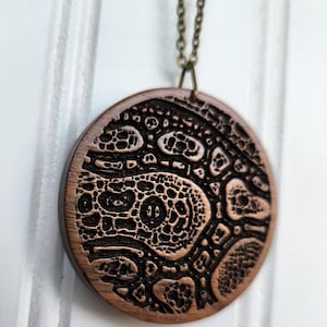 May include: A round, wooden pendant necklace with a detailed, carved design. The pendant is a warm brown color with a dark, intricate pattern. The necklace chain is a bronze color.