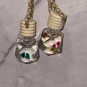 Op de afbeelding: Twee kleine, doorzichtige, geometrische glazen flesjes gevuld met vloeistof en gedroogde bloemen, elk voorzien van een geribbelde houten dop en opgehangen aan een gevlochten beige en goudkleurig koord. Het ene flesje bevat blauwgroene bloemen, het andere roze en gele bloemen.