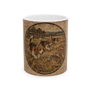 May include: A ceramic mug with a white interior and a tan, wood-grain textured exterior. The front features an oval-framed illustration of three tricolor beagle dogs running through a grassy field, chasing two brown rabbits in a rural landscape.