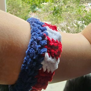 May include: A crocheted bracelet in red, white, and blue. The bracelet is made of a thick, textured yarn and is wrapped around an arm. The colors are arranged in a striped pattern, with blue on the top and bottom and red and white in the middle.