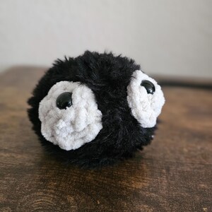 May include: A handmade plush toy with a round shape, featuring black fur and two large, white, fluffy eye-like shapes with black button centers. The toy is resting on a wooden surface.