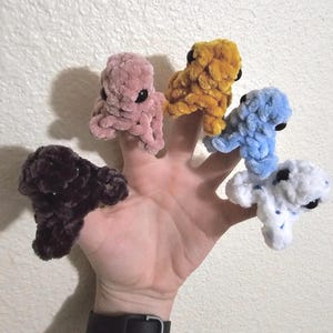 May include: Five colorful, crocheted octopus finger puppets. The puppets are in shades of pink, yellow, blue, white, and dark brown. Each octopus has black button eyes. The puppets are displayed on a hand.