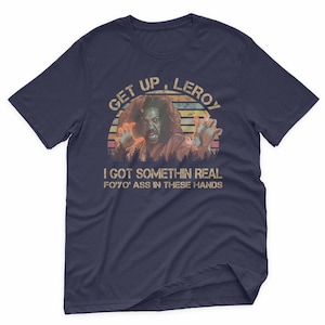 May include: Navy blue t-shirt with a distressed graphic featuring a person with outstretched hands and the text "GET UP, LEROY" above and "I GOT SOMETHIN REAL FO'YO' ASS IN THESE HANDS" below. The graphic has a vintage, retro style.