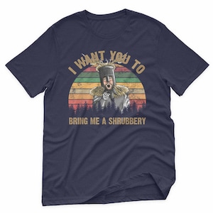 May include: Navy blue t-shirt with a retro design featuring a knight and the text "I WANT YOU TO BRING ME A SHRUBBERY". The graphic includes a sunset and forest silhouette.