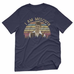 May include: Navy blue t-shirt with the text "I AM MCLOVIN" in a vintage style. The graphic features a portrait of a man with glasses, set against a sunset design with trees at the bottom. A fun, pop culture themed tee.