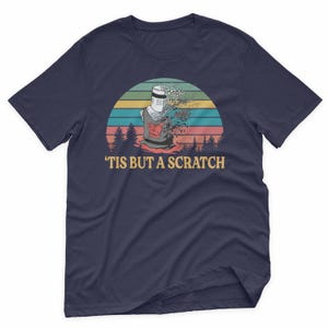 May include: Navy blue t-shirt featuring a graphic of a knight with the text "'TIS BUT A SCRATCH" in a retro sunset design. The knight is depicted with a missing arm and head, with a red emblem on the chest.