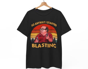May include: Black t-shirt featuring a graphic of a man holding two guns, set against a sunset backdrop. The text "SO ANYWAY I STARTED BLASTING" is printed above and below the image. The shirt is hanging on a wooden hanger.