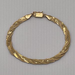 Könnte beinhalten: Ein goldfarbenes Armband mit einem geflochtenen, mehrsträngigen Design und einer texturierten, reflektierenden Oberfläche. Das Schmuckstück wird durch einen rechteckigen Kastenschloss-Verschluss oben gesichert, der die beiden Enden verbindet.