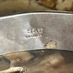 May include: Close-up of a silver-toned metal object with the engraved text "S&E GUARDIAN". The object has a reflective surface and appears to be part of a larger piece. The background is a light brown color.