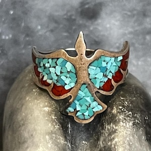 Op de afbeelding: Een zilveren ring met een vogelvormig inlegontwerp. De vogel is gevuld met kleine, verbrijzelde turkoois steenfragmenten in het midden en de vleugels, geaccentueerd door rode steensegmenten langs de buitenranden van de vleugels en staartveren.