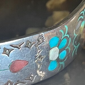 May include: Close-up of a silver bracelet with intricate floral and geometric designs. The bracelet features inlaid turquoise, white, and red accents. The design includes a red tulip and a stylized flower with turquoise petals.